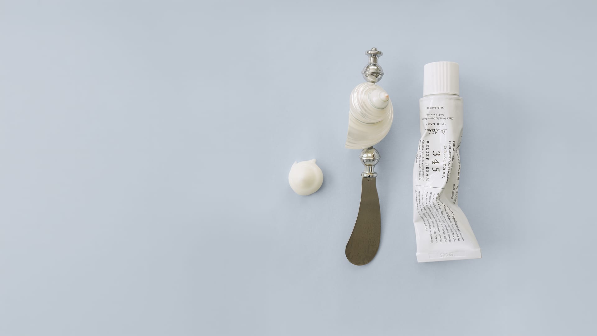 Tubo de crema, espátula y una concha con un poco de crema sobre fondo gris azulado claro